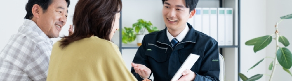 「塗装業者の担当者が笑顔で相談に応じる様子、テーブル越しに中年夫婦と話し合いをしている室内のシーン」