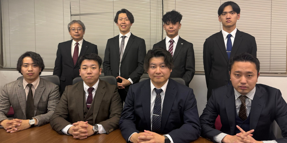 会議室でスーツを着た男性社員8名が前列・後列に分かれて整列し、カメラに向かって正面から写る集合写真。
