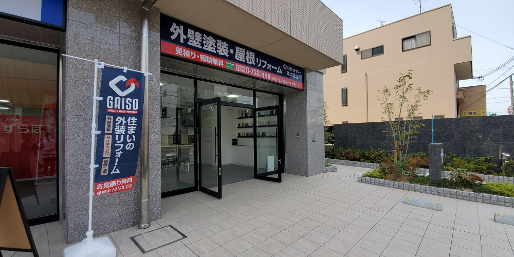 ガイソー東大阪店の店舗外観-ガラス扉のショールーム入口と「外壁塗装・屋根リフォーム」の看板、店頭に立つ青いのぼり旗