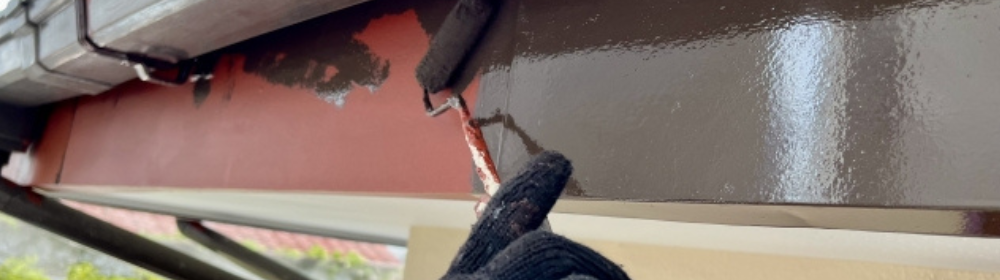 「建物の鉄部にローラーで塗装を施す作業中の手元、黒い手袋をした職人が赤茶色の下地にこげ茶色を塗っている場面」