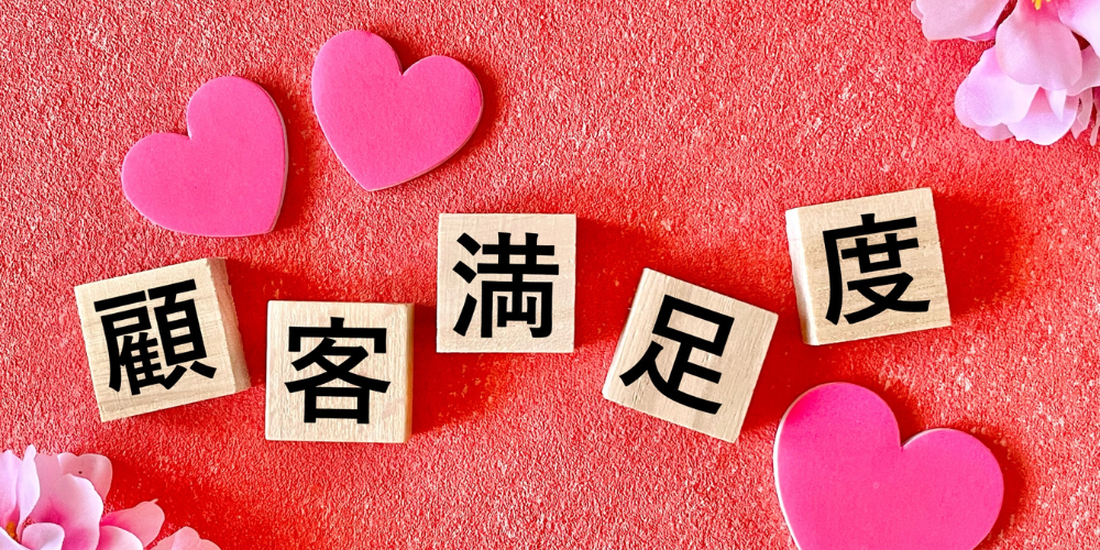 「顧客満足度」の文字が書かれた木のブロックと、ピンクのハートと桜の装飾が並ぶ赤背景の画像