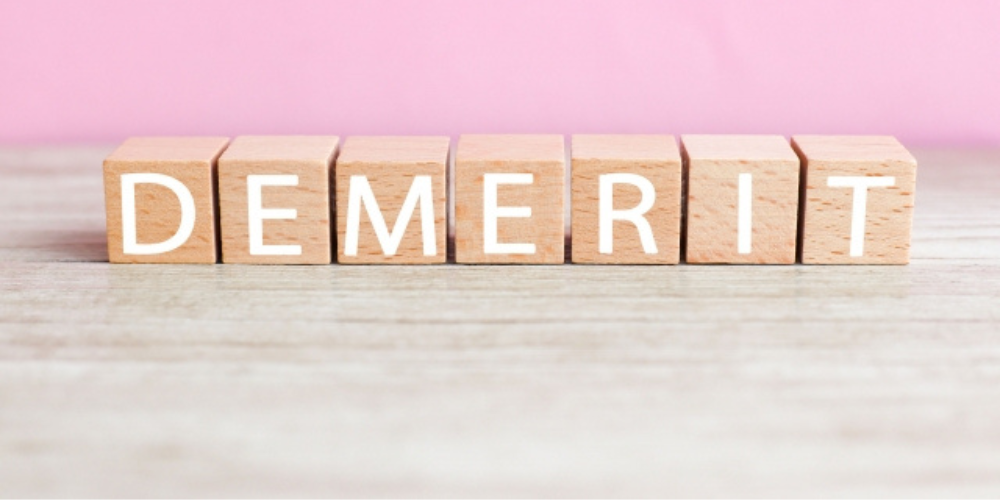 木製ブロックに「DEMERIT」と白い文字が並び、背景はピンク色。