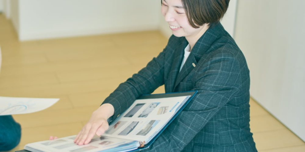 スーツ姿の女性スタッフが室内で施工写真が綴じられたバインダーを広げ、笑顔で説明しているシーン。