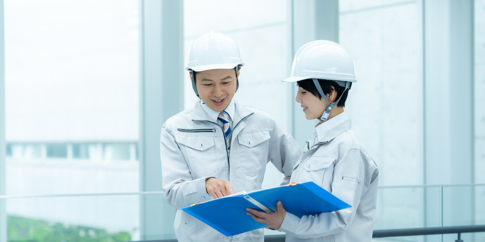 白い作業服とヘルメットを着用した男女が青いファイルを見ながら相談している様子