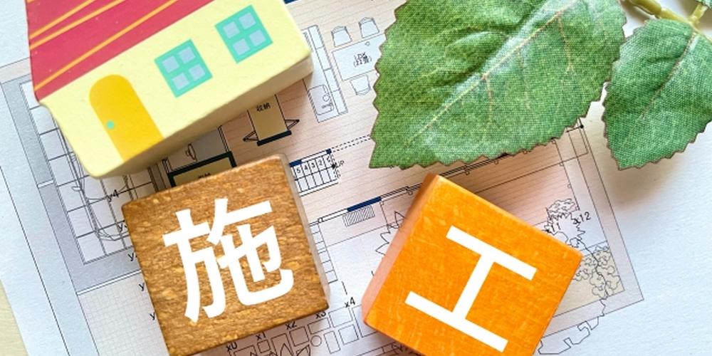 設計図の上に置かれた家の模型と、「施工」の漢字が書かれた木製ブロック、フェイクグリーンの葉が添えられているイメージ