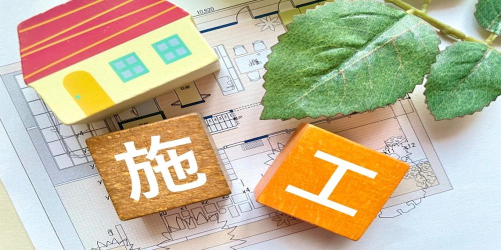 「施工」の文字が書かれたブロックと家の模型、設計図、葉の飾りが並ぶイメージ写真。