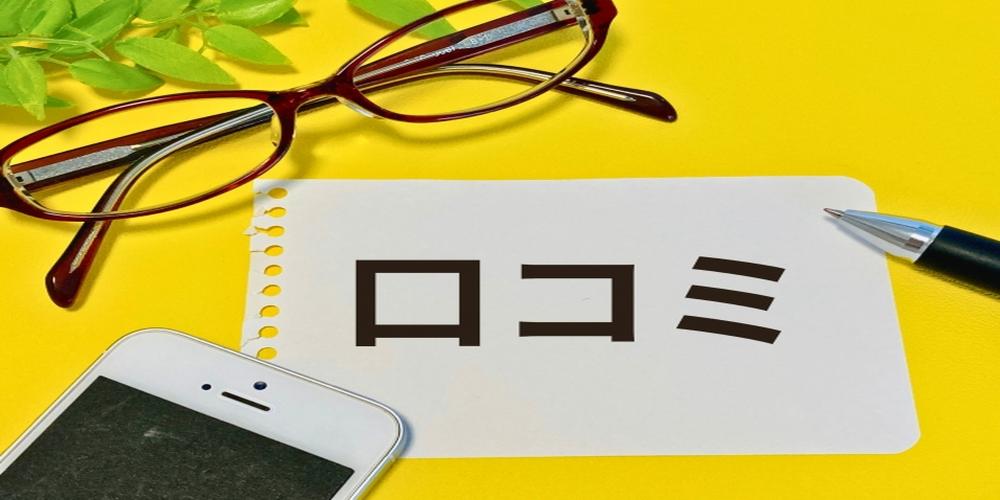 「口コミ」と書かれたメモとスマートフォン、メガネ、ペンが並ぶイメージ。背景は明るい黄色。
