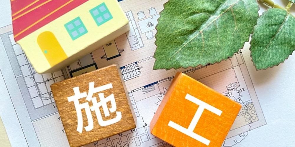 「住宅設計図の上に配置された『施工』の文字が書かれた積み木と家の模型、そして緑の葉」