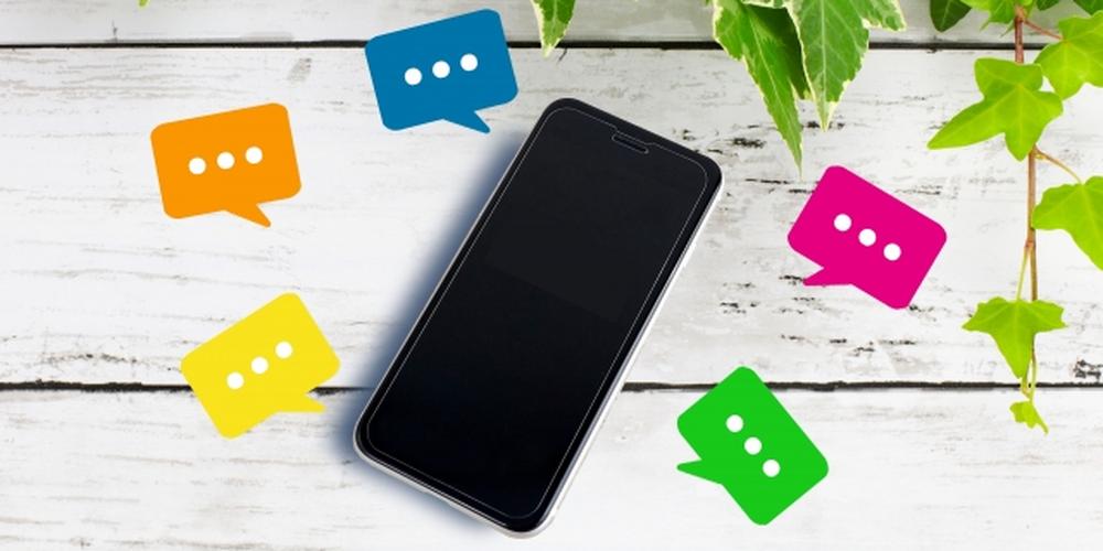 「木目の背景に置かれたスマートフォンと、周囲に浮かぶカラフルな吹き出しアイコンと緑のアイビー」
