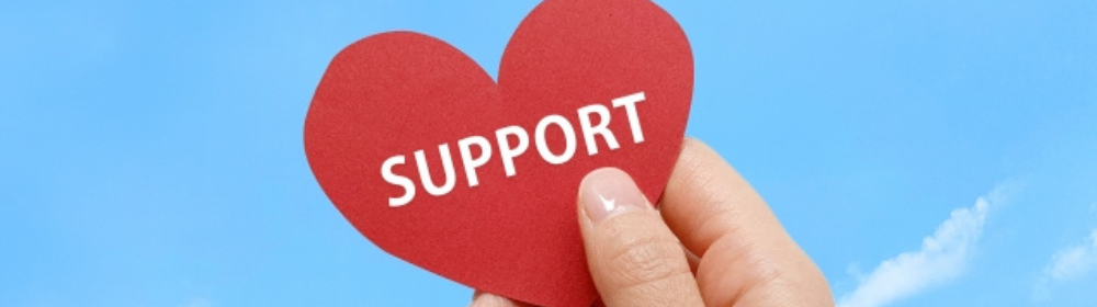 青空を背景に、赤いハート型のカードに「SUPPORT」と書かれたものを手で持っている。