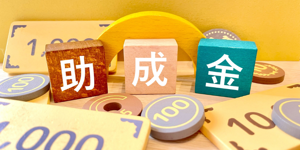 「助成金」という漢字の積み木と、おもちゃのお金が置かれている