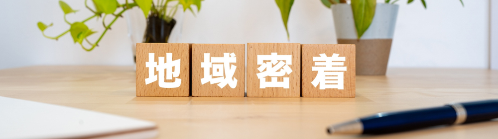 木製ブロックに「地域密着」と書かれ、観葉植物とペンが置かれたデスク