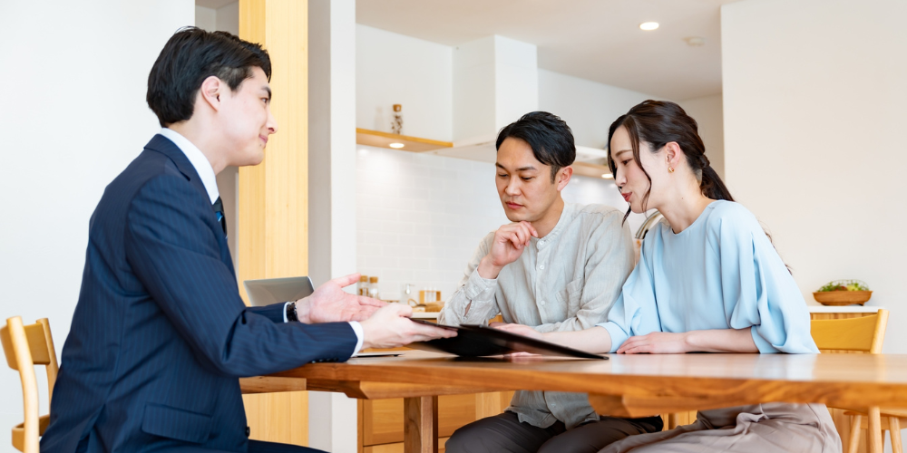 自宅のリビングで、スーツ姿の担当者から説明を受ける若い夫婦