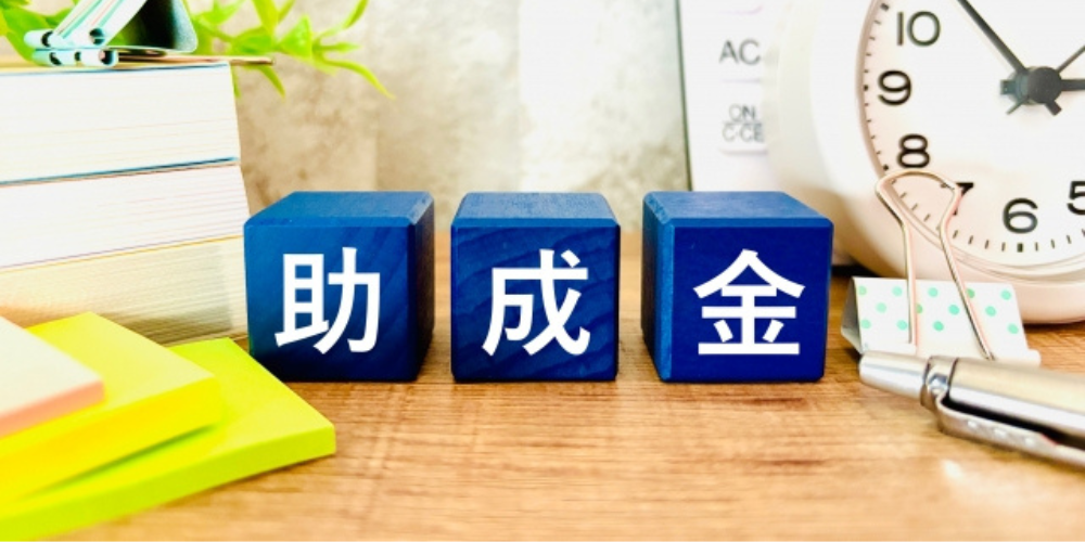 「助成金」と書かれた青い積み木が並ぶ机の上のイメージ、時計や文房具が背景に配置