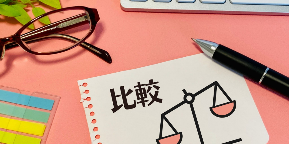 ピンクの作業スペースで「比較」と秤のイラストが描かれたメモ用紙とメガネ・ペン・付箋