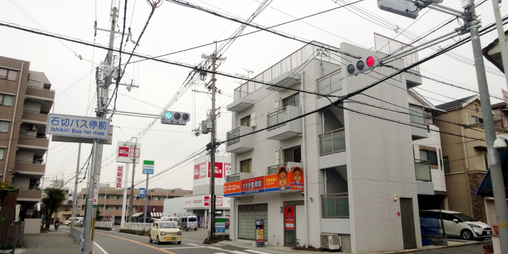 「石切バス停前」標識と電線が張り巡らされた交差点、低層マンションが並ぶ街並み