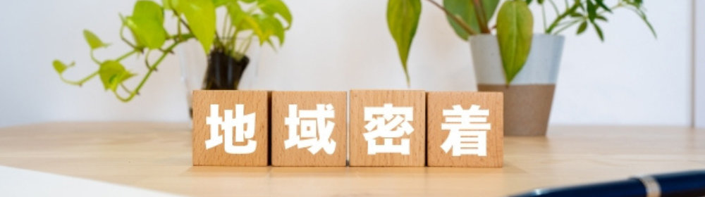 木のブロックに書かれた「地域密着」の文字と観葉植物の背景。