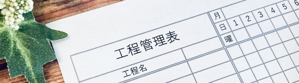 「工程管理表」の用紙とペン、キーボードと植物が置かれたデスク