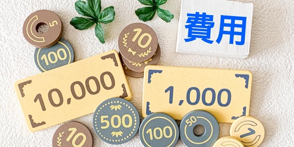 費用をイメージした木製のコインや紙幣が並ぶ背景に、「費用」と書かれた木札が置かれている写真。家計や支出管理、費用に関する話題に適したイメージ。