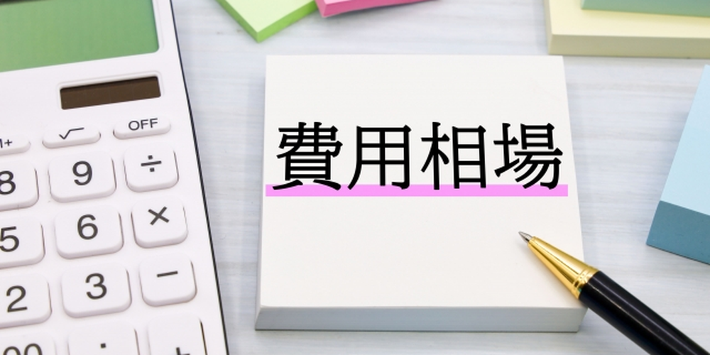 「費用相場」と書かれたメモ用紙と電卓、ペン、カラフルな付箋が並ぶコスト計算のイメージ画像