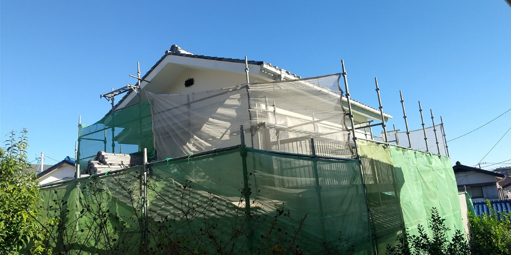 青空の下、足場と養生ネットで囲まれて外壁塗装工事中の戸建て住宅