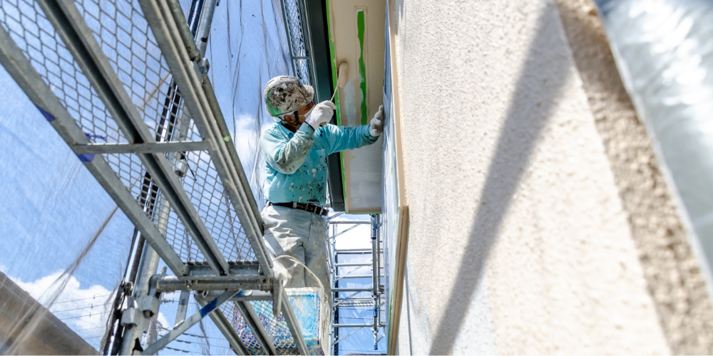 外壁の足場に立ち、ヘルメットと作業着で刷毛塗りを行う塗装職人の作業風景