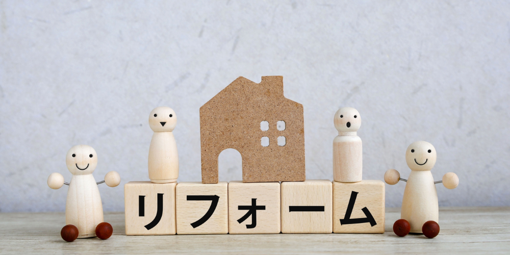 リフォームを象徴する木製の家とブロック、両脇に笑顔と驚きの表情をしたフィギュアが並んでいる