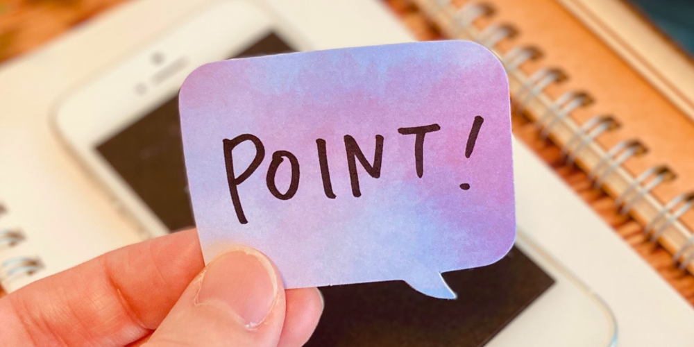 ノートやスマートフォンの上で、手が紫ブルーの吹き出し型付箋に「POINT!」と手書きされている注目ポイント提示イメージ