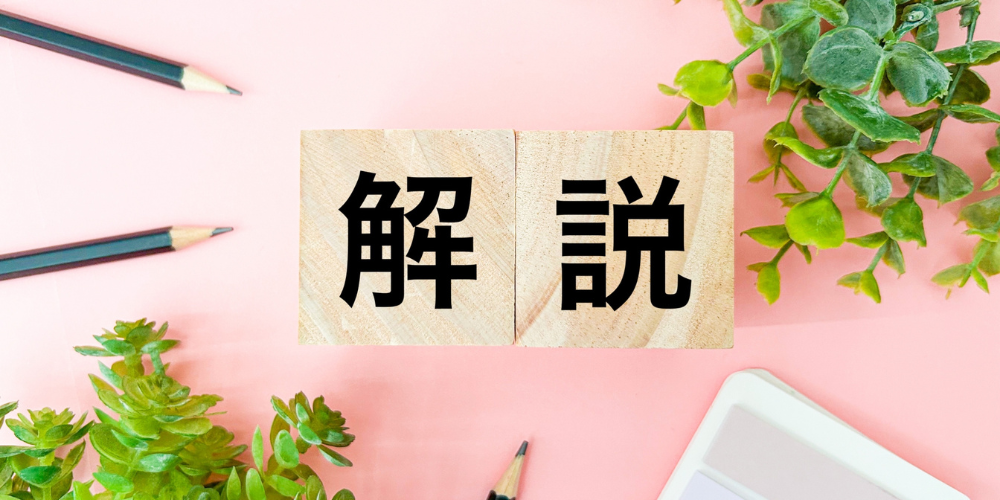 ピンクのデスク上に鉛筆やグリーンの観葉植物が配置され、木製ブロックに「解」「説」と黒字で書かれた解説強調イメージ