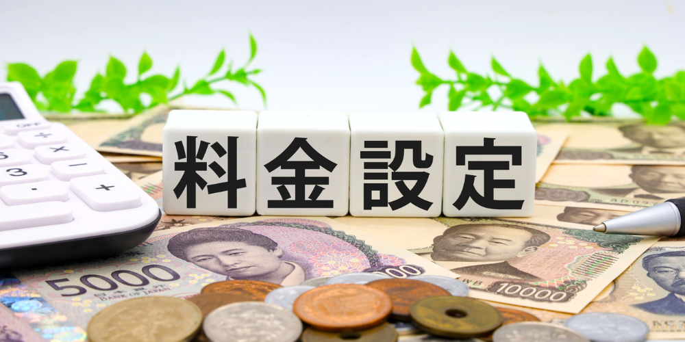 白い電卓、千円札や一万円札、硬貨を散らした木目デスク上に、白いキューブに黒字で「料金設定」と書かれたコスト表示イメージ