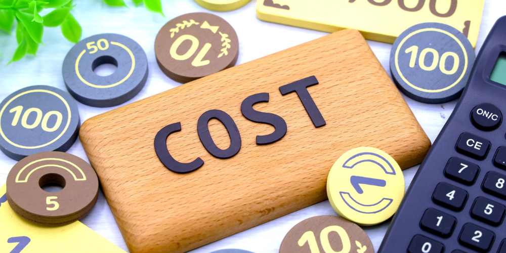 木製の板に黒い大文字で「COST」と書かれ、その周囲に円形のコイン風オブジェや電卓が配置されたコストイメージ