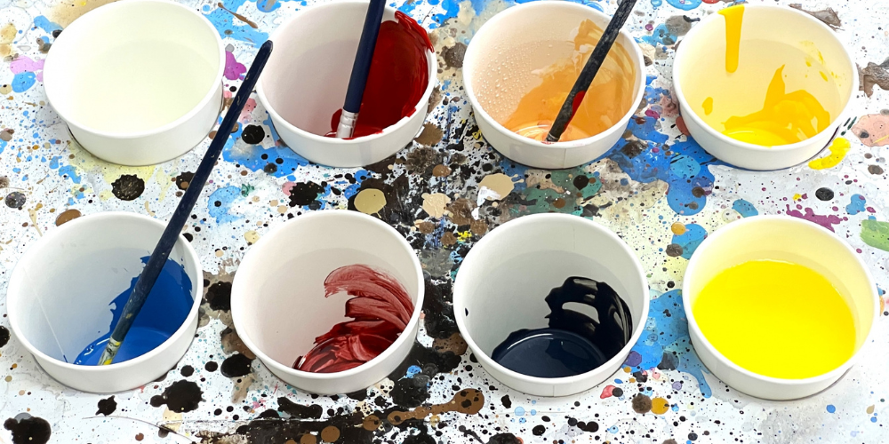 ペンキ入れの紙コップに赤・オレンジ・黄色・青・黒などの絵の具が入っており、筆が差し込まれたまま散らかった作業台の上に並ぶ塗装材料イメージ
