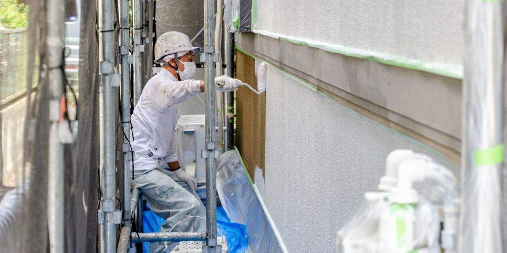 足場に囲まれた建物外壁で、ヘルメットとマスクを着用した職人がローラーで塗装作業を行っている様子