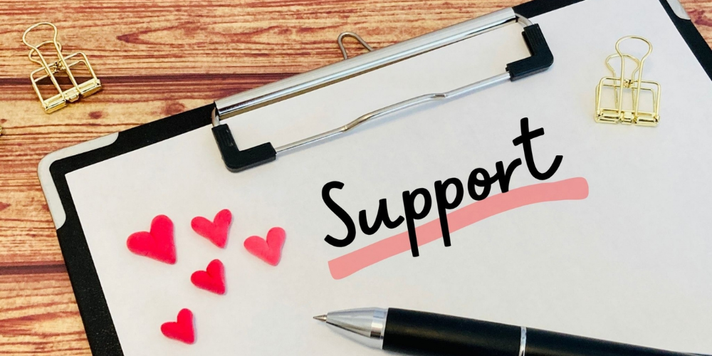 木目のデスクに置かれたクリップボードの紙面に英字で「Support」、その横に赤いハート形ステッカーが散りばめられたサポート強調イメージ