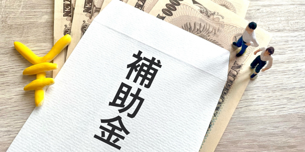 複数の一万円札と五千円札を重ね、その上に白い封筒風ボードで「補助金」と黒字で書かれ、横に小さな木製フィギュア2体が立つ補助金案内イメージ