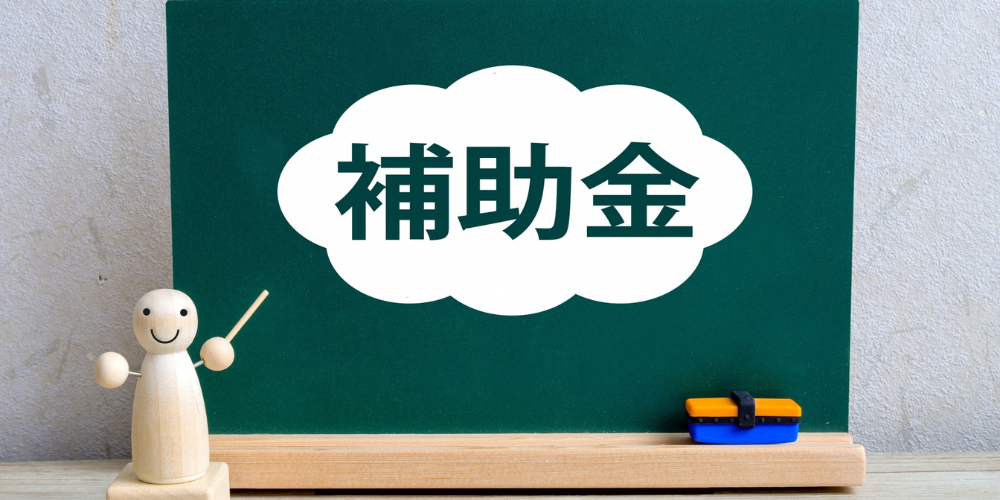 黒板の前に木製フィギュアが立ち、黒板中央の白い吹き出しに濃緑字で「補助金」と書かれた補助金案内イメージ